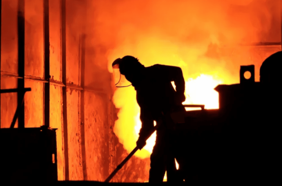 Worker silhouetted against intense fiery furnace light.